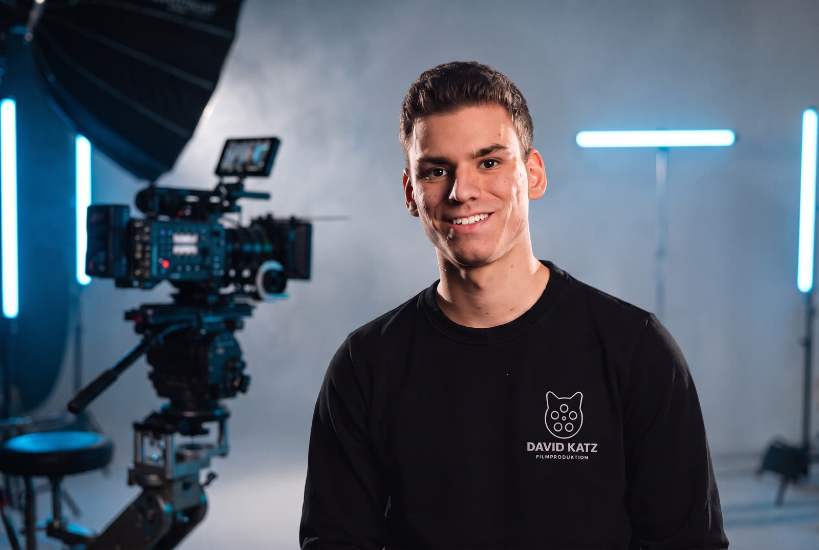 David Katz lächelt in die Kamera, hinter ihm ein Filmset mit Kinokamera, Softbox und blauem Licht.